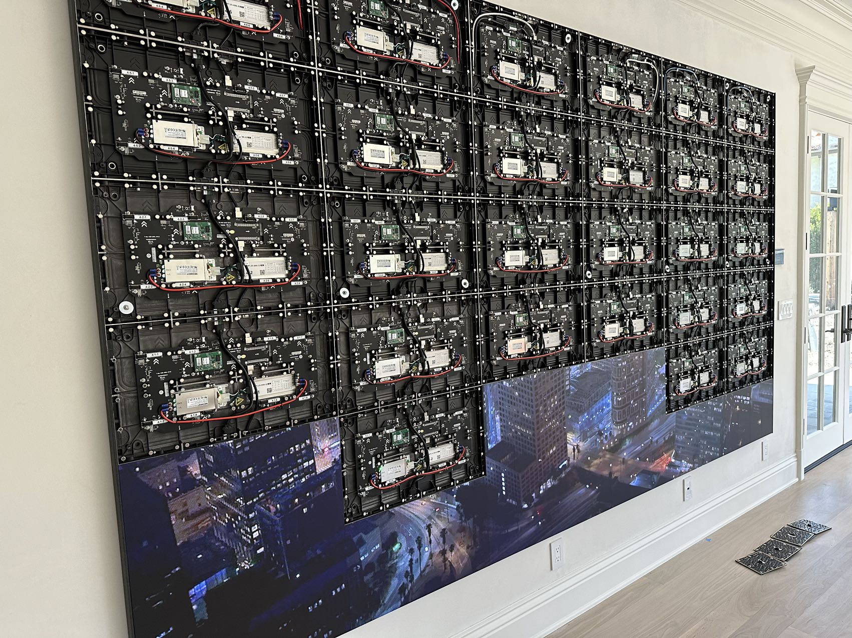 Home LED wall mid-installation with front modules removed, exposing receiving cards, power supplies and hub boards on the cabinet backs while the lower rows play content