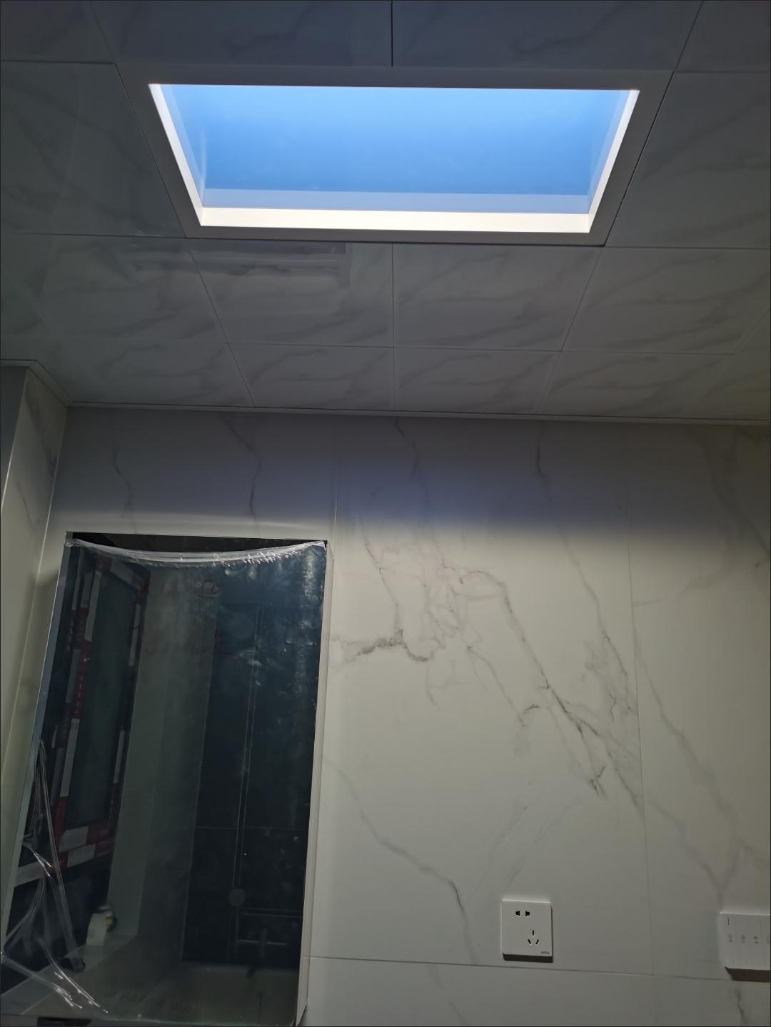 LED skylight panel in the ceiling of a windowless bathroom with marble tile walls, casting daylight down the wall