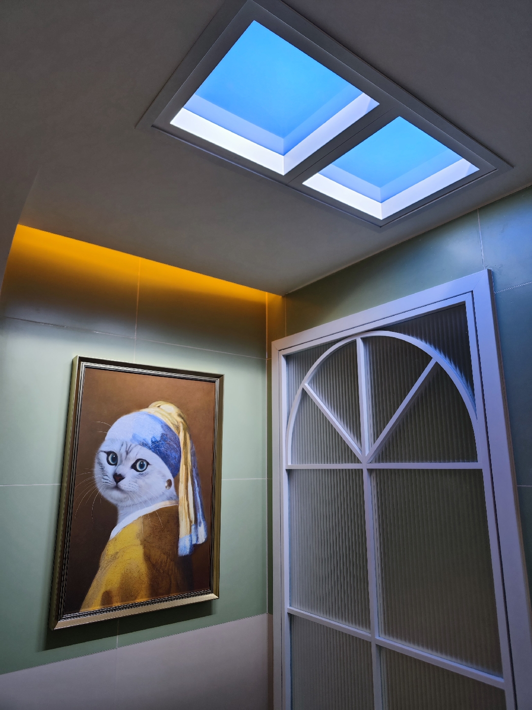 Two LED skylight panels installed side by side in the ceiling of a windowless entryway, showing a bright blue sky above a warm cove light and a green-panelled wall